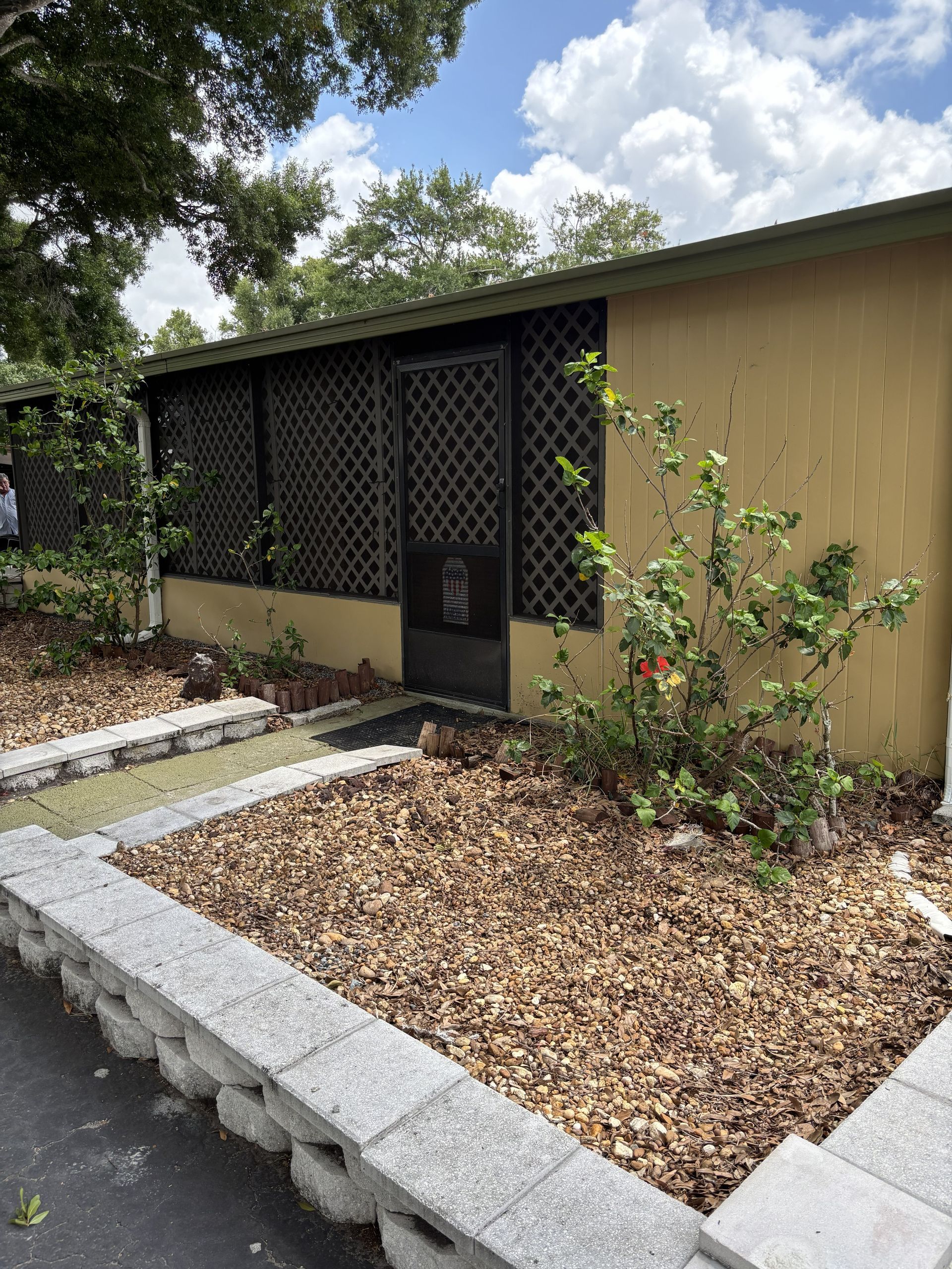 Mobile home with black lattice screen, front door, and landscaped gravel beds bordered by concrete.