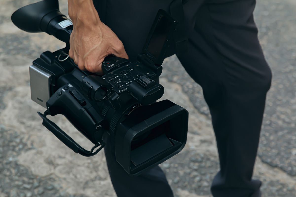 Person carrying a black video camera by its handle on a street.