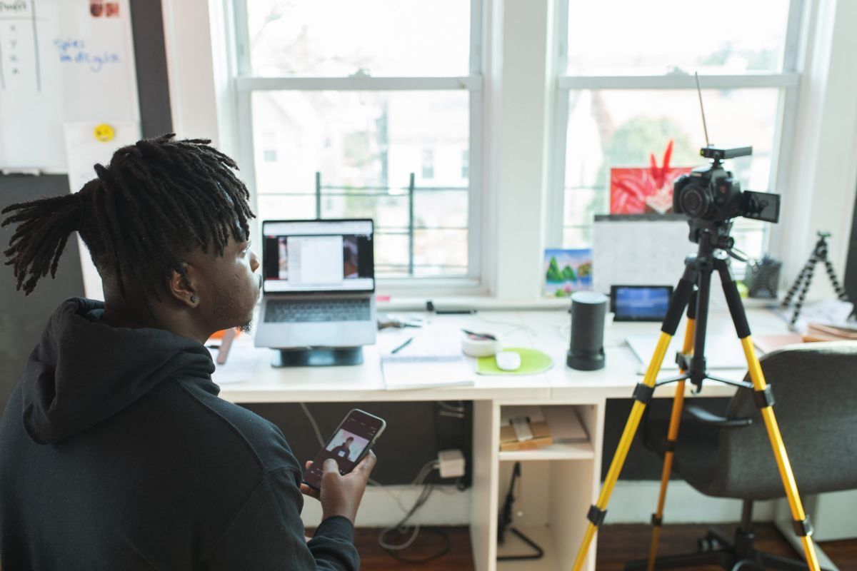 Person holding a phone beside a tripod-mounted camera in a bright room with large windows