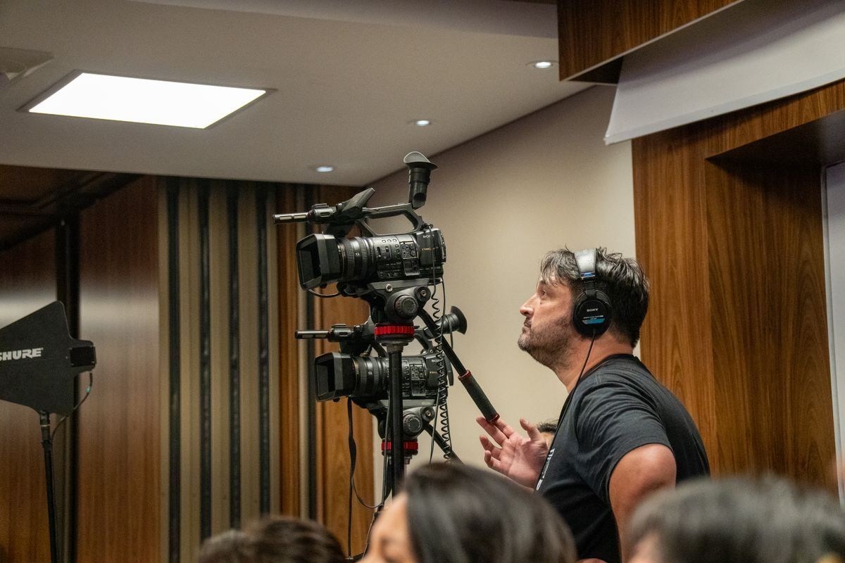 Person wearing headphones films with a large camera in a wood-paneled room during an event.