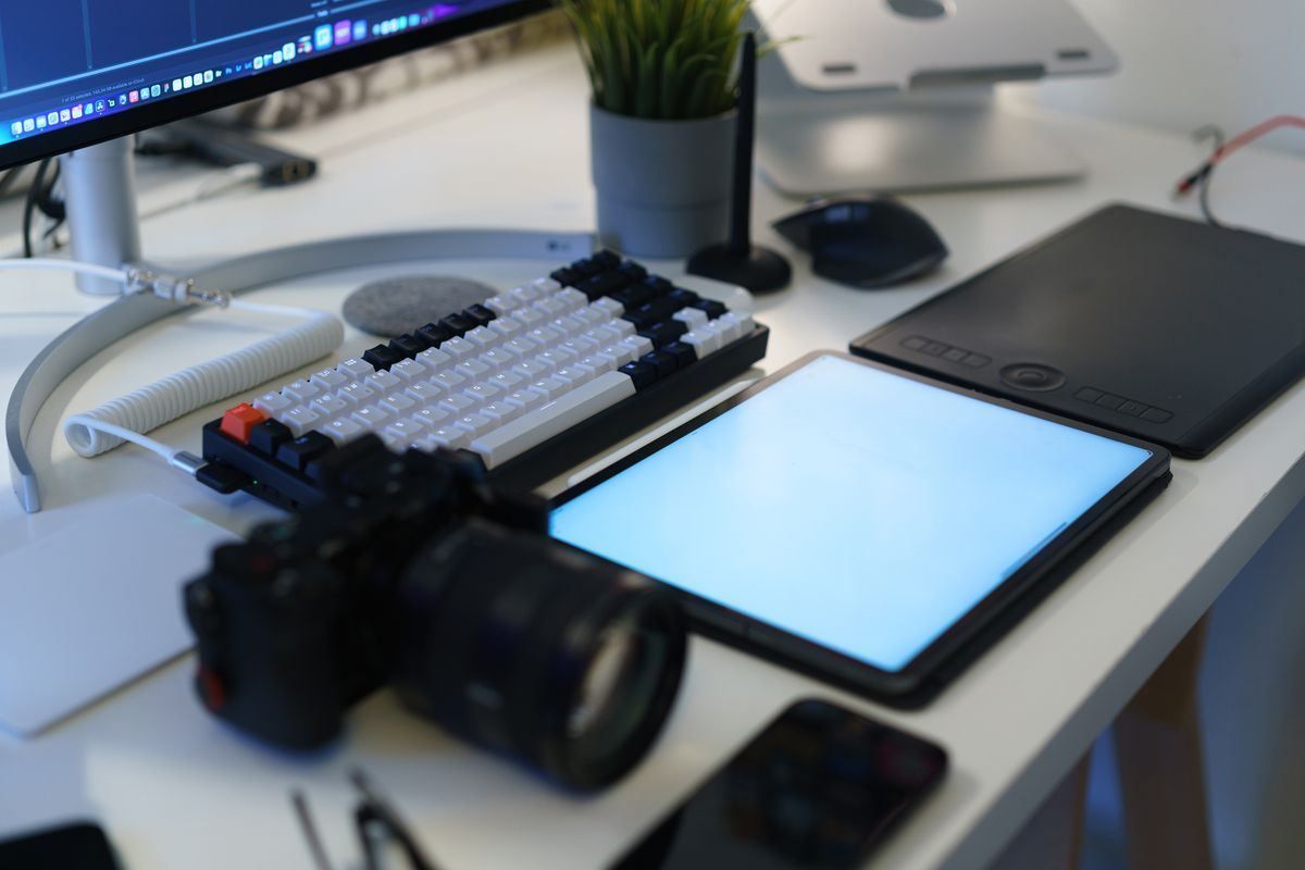 Modern desk setup with monitor, keyboard, tablet, camera, and notebook on a white desk