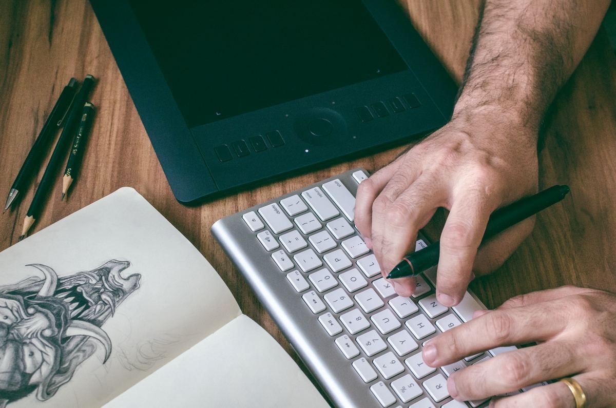 Hands typing on a keyboard beside a sketchbook and drawing tablet on a wooden desk
