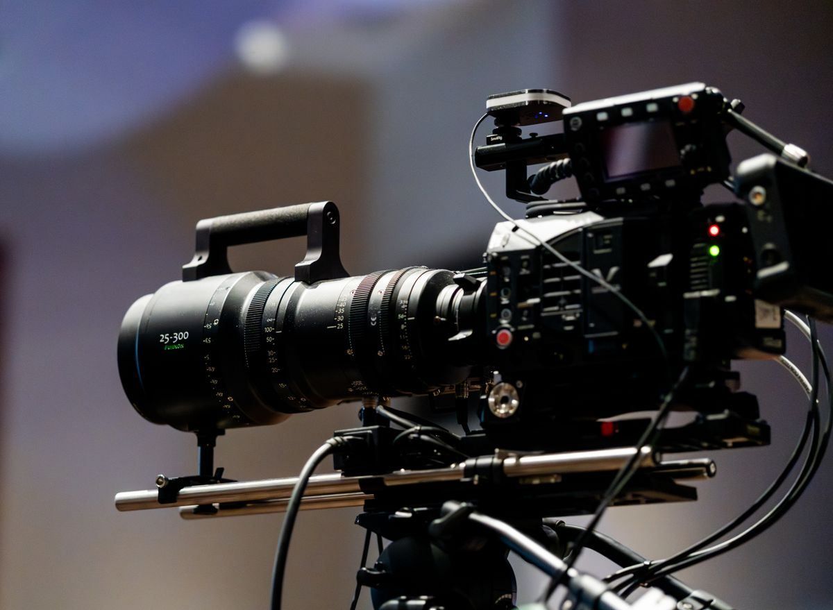 Professional video camera on a tripod with a large lens, set against a blurred indoor background