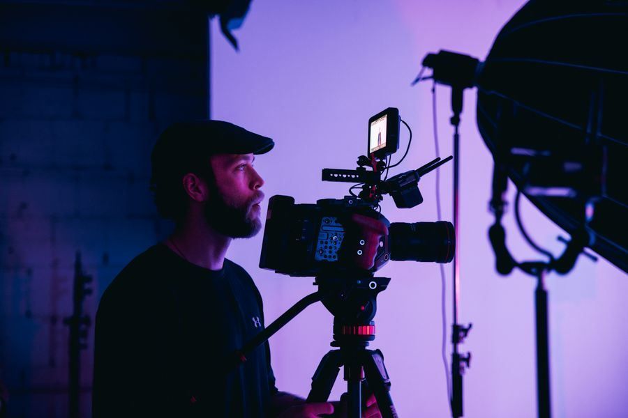 Videographer filming with a camera on tripod in purple-lit studio, adjusting monitor and lens
