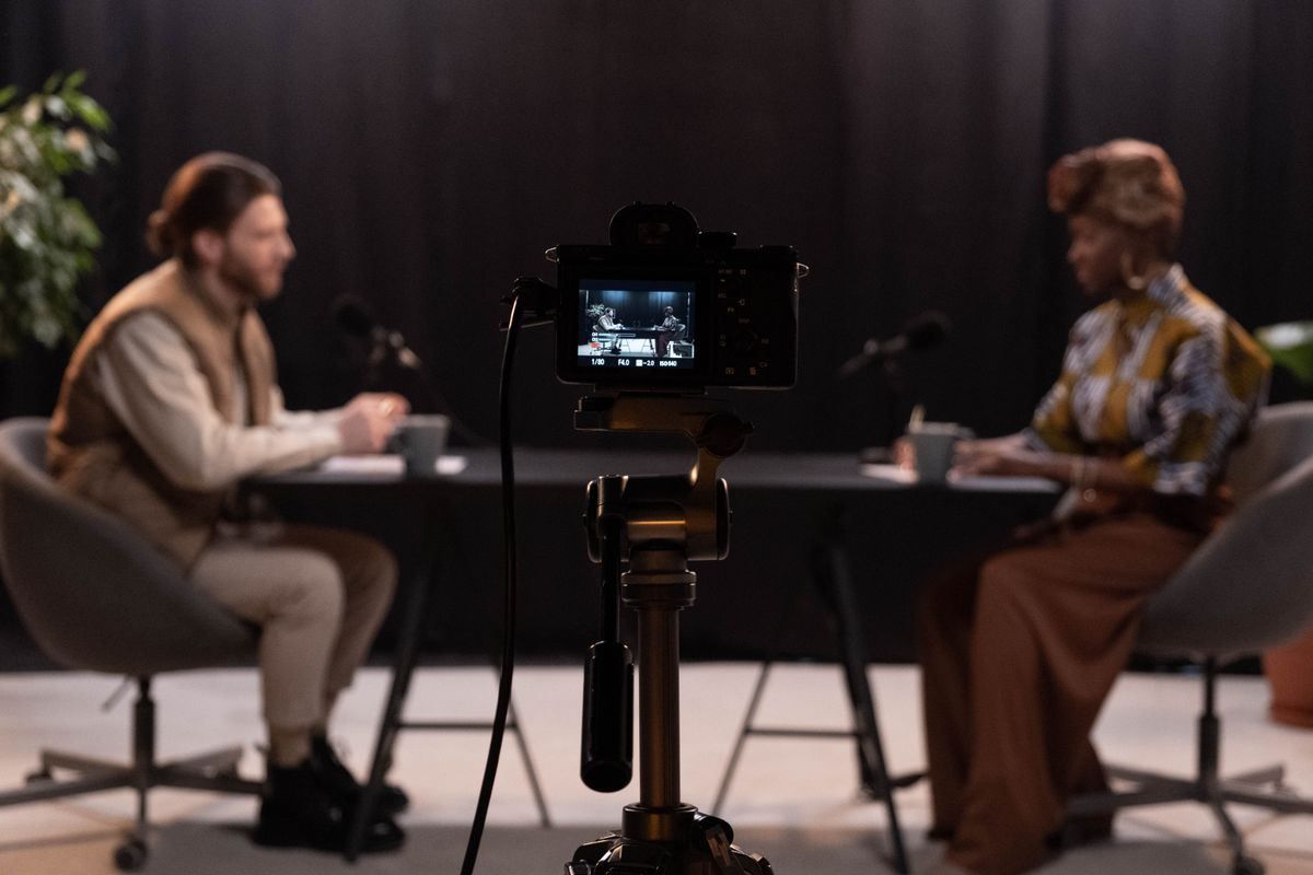 Two people seated at a table during a filmed interview, seen through a camera on a tripod.