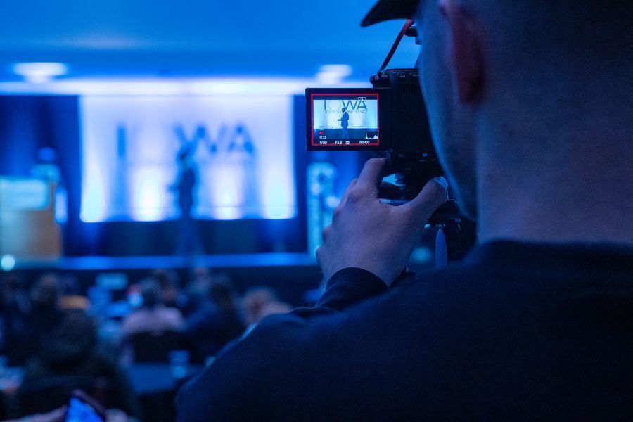 Person filming a blue-lit conference stage with a camcorder from the audience