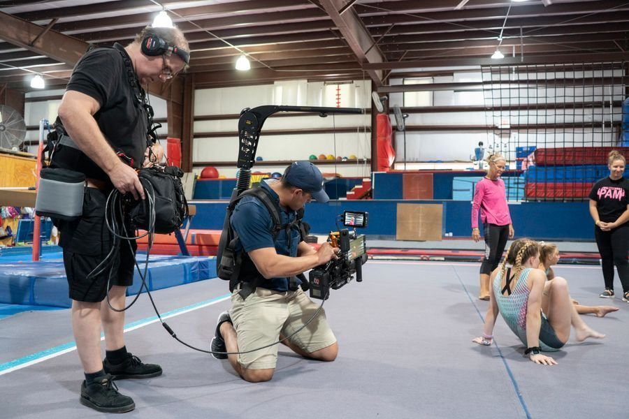 Two camera operators film gymnasts on mats inside a large indoor gymnasium