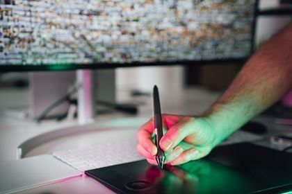 Hand drawing on a tablet beside a monitor, lit by pink and green light