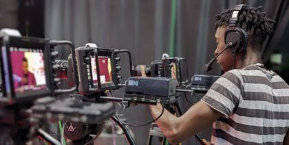 Camera operator filming with multiple monitors and a headset in a studio setting