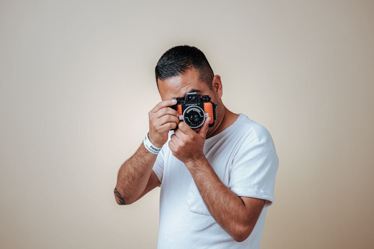 Person in a white shirt holding a camera and taking a photo against a plain wall.