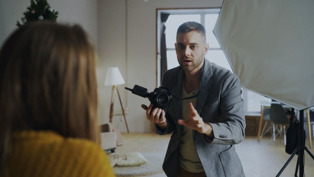 Photographer holding a camera and gesturing during a portrait shoot in a studio, with a model in the foreground