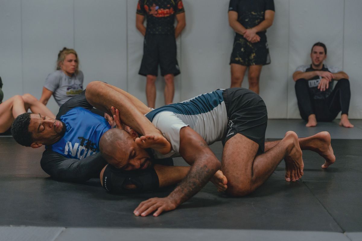 Two wrestlers grappling on a mat in a gym, with spectators sitting nearby.