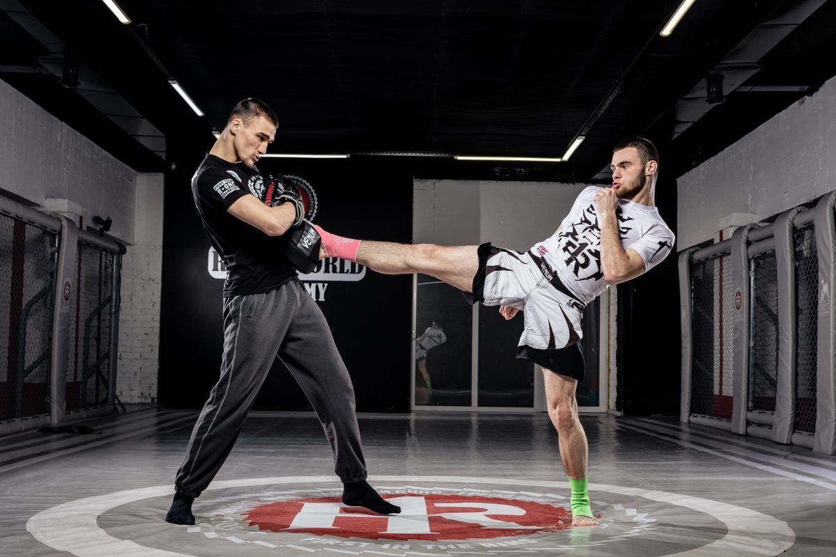 Two men sparring in a gym, one kicking the other’s torso on a mat.