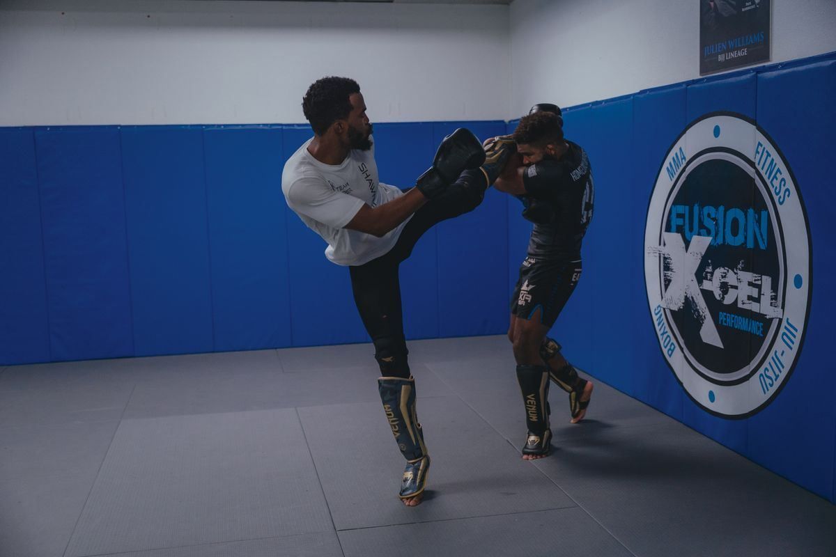 Two martial artists sparring in a blue gym, one delivering a high kick with padded gear on both.