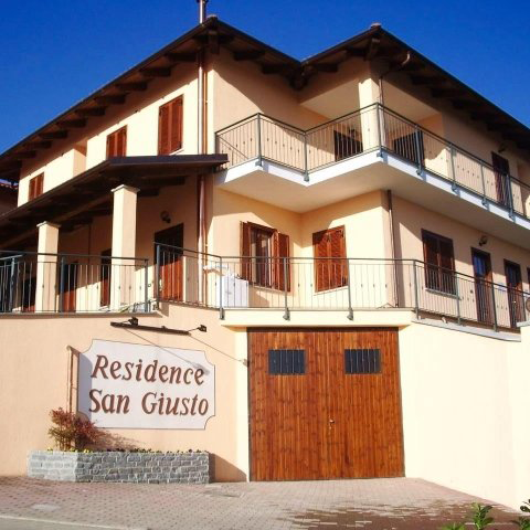 Edificio del Residence San Giusto con pareti beige, persiane marroni, balconi e un portone da garage in legno sotto un cielo azzurro.
