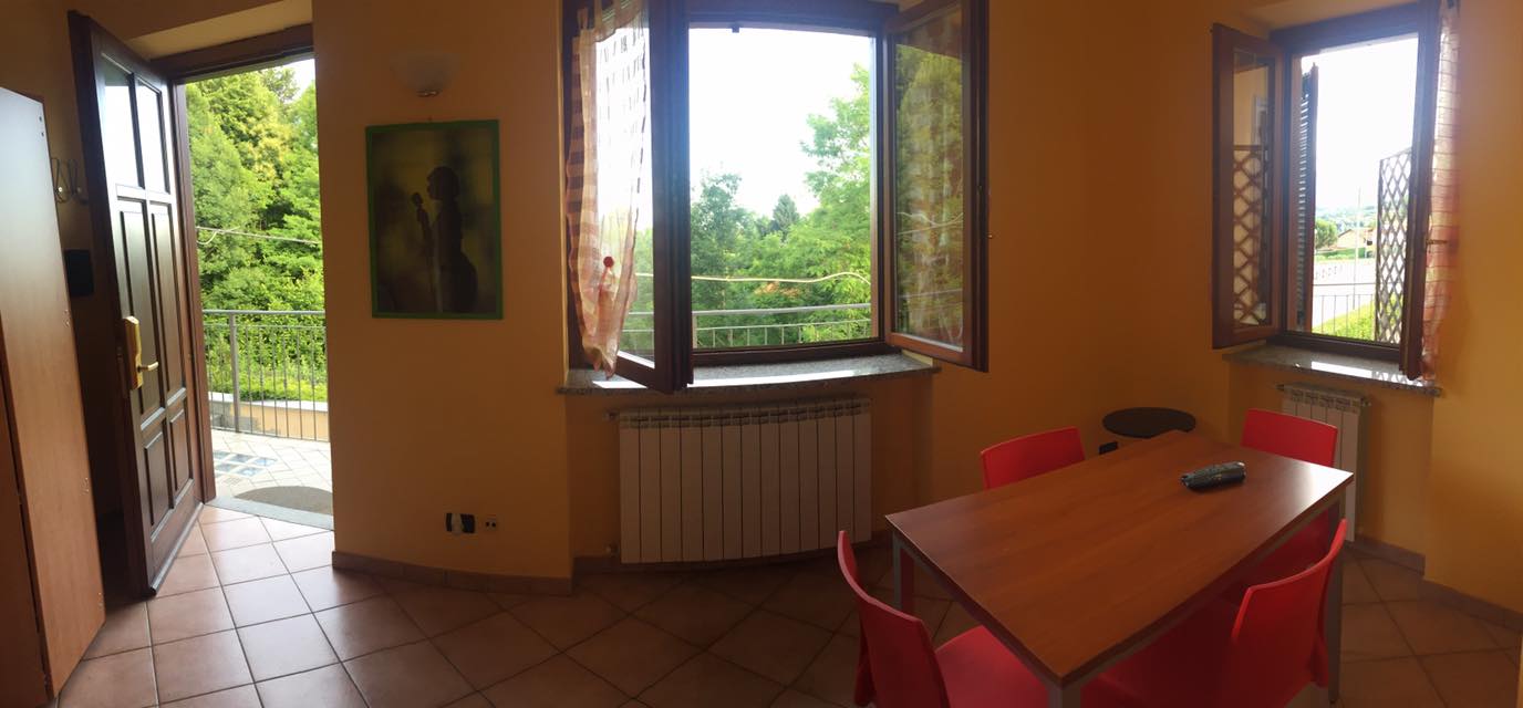 Interno di una stanza con un tavolo e sedie rosse, una porta aperta su un balcone e grandi finestre.