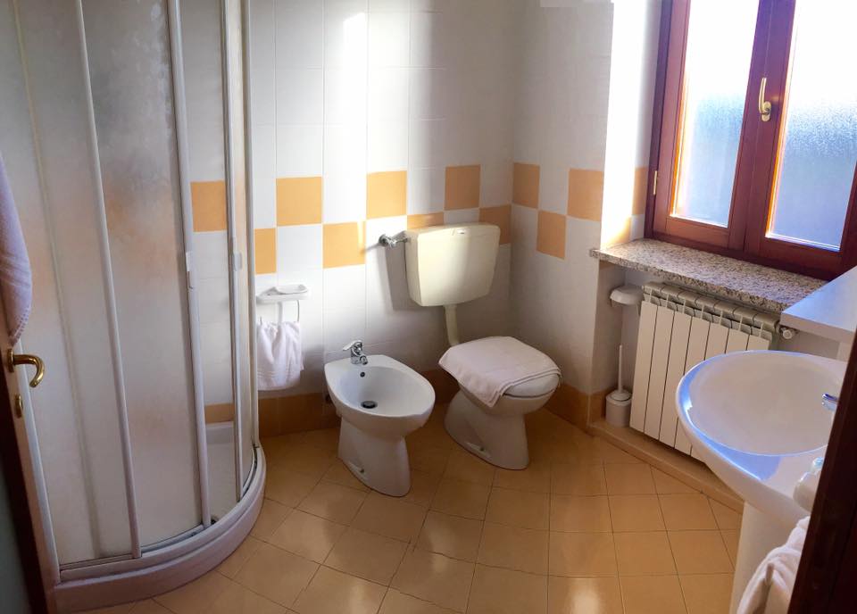 Bagno con doccia, WC, bidet e lavandino. Piastrelle gialle e bianche e pavimento color cuoio. La luce del sole filtra a fiumi.