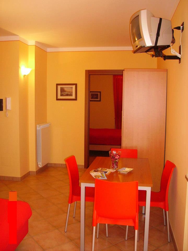 Zona pranzo con sedie e tavolo arancioni, TV montata a parete, porta che conduce alla stanza rossa.