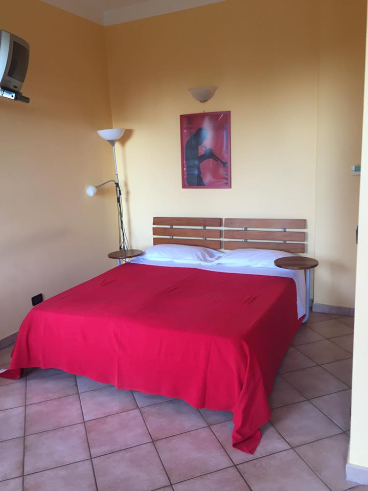 Camera da letto con copriletto rosso, testiera in legno, lampada da terra e opere d'arte; pareti gialle e pavimento piastrellato.