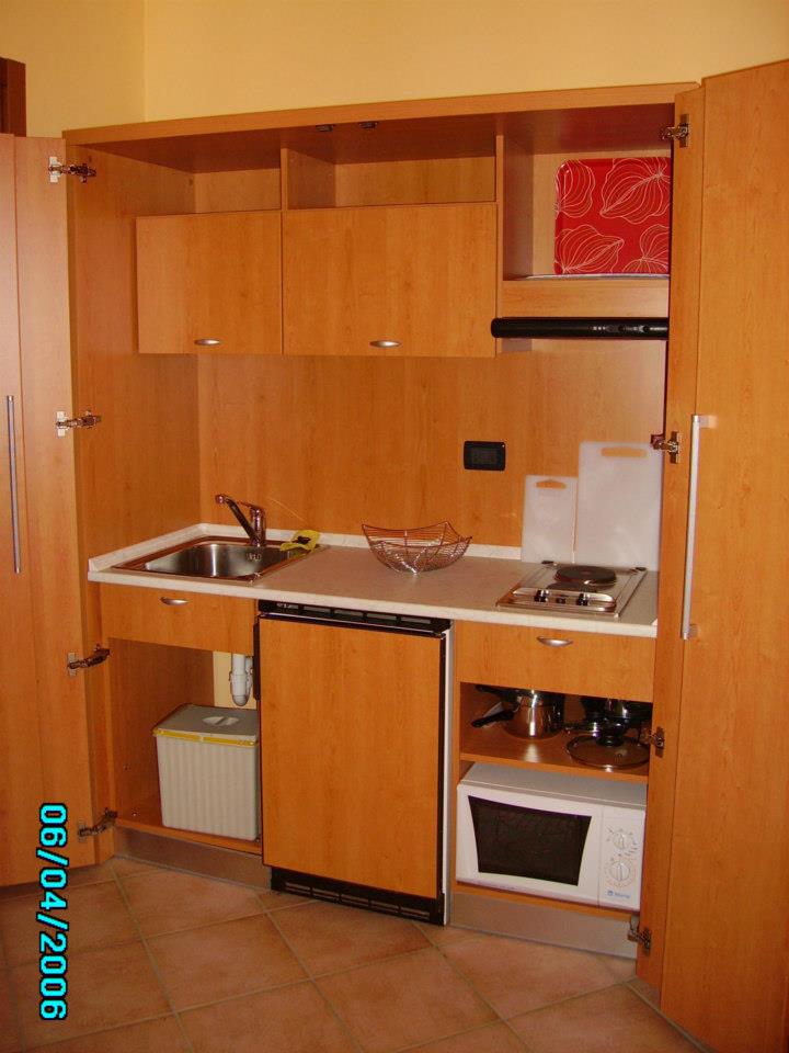 Piccola cucina in legno con elettrodomestici, tra cui forno a microonde, frigorifero e fornello.