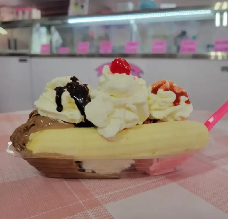 Banana split dessert with whipped cream, chocolate syrup, cherry, and strawberry topping in a pink diner setting