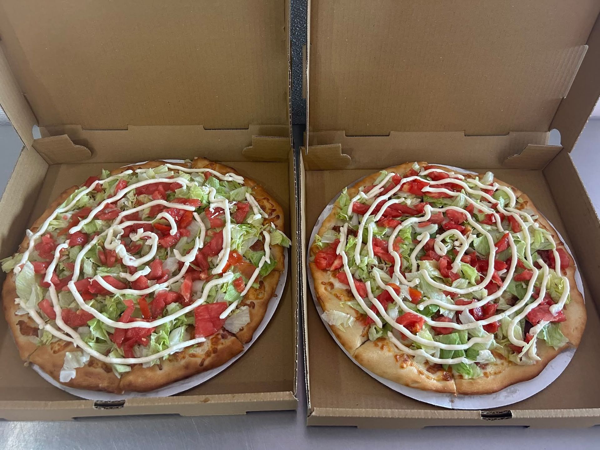 Two pizzas in open boxes, topped with white sauce drizzles and green vegetables.