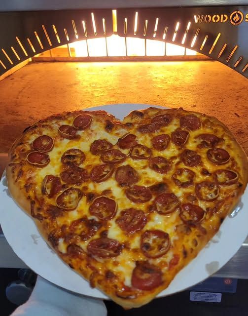 Heart-shaped pepperoni pizza on a white tray inside a glowing wood-fired oven.
