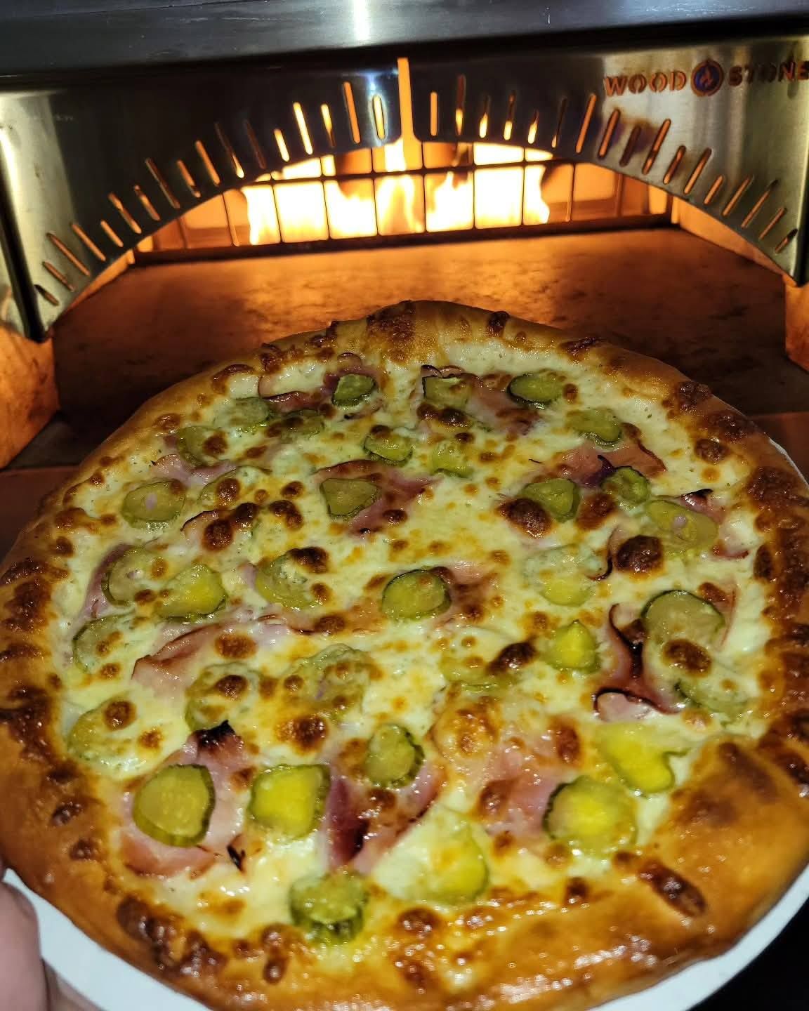 Pizza baking in a wood-fired oven, topped with melted cheese, green peppers, and olives