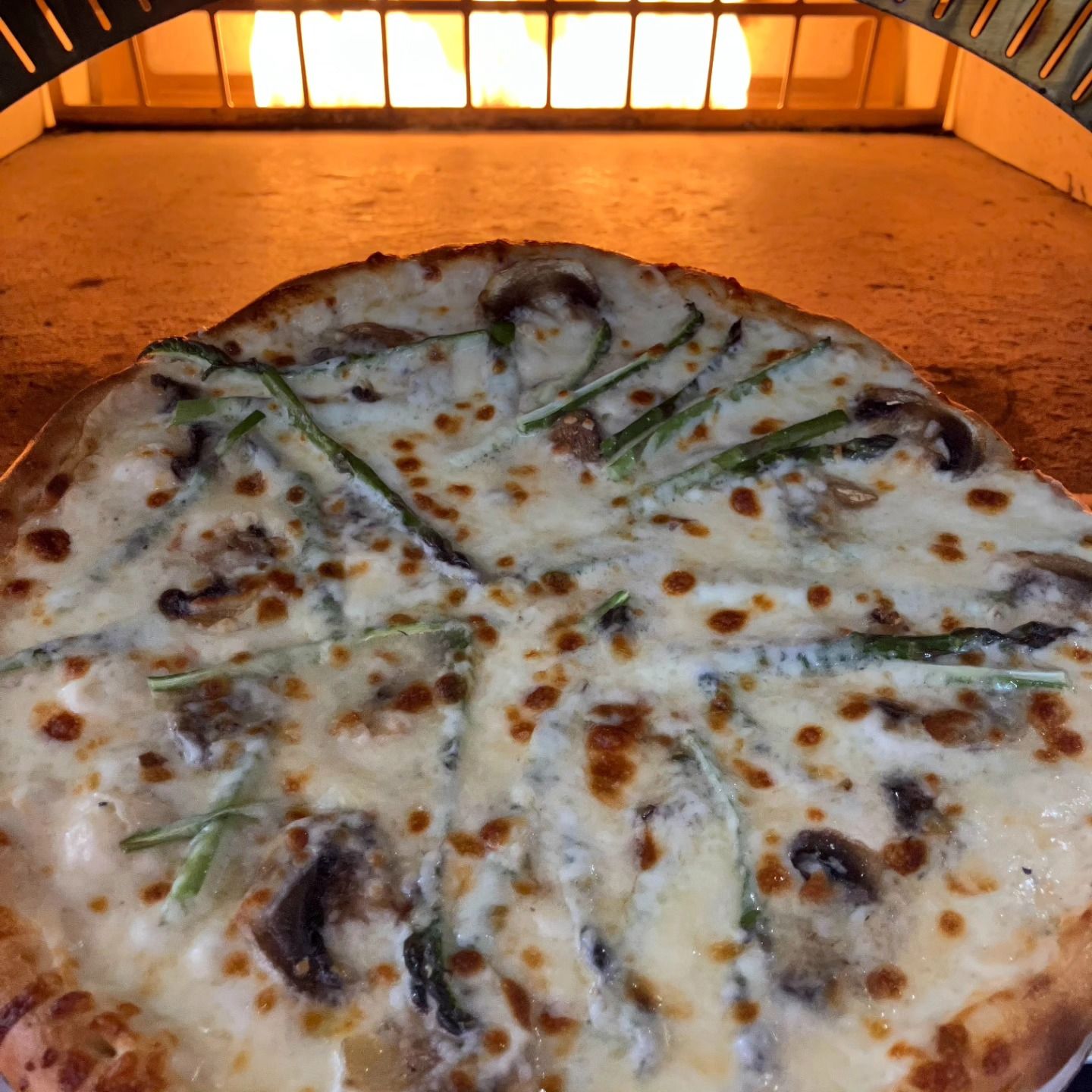 Cheese and mushroom pizza baking inside a wood-fired oven, lit by warm orange flames.