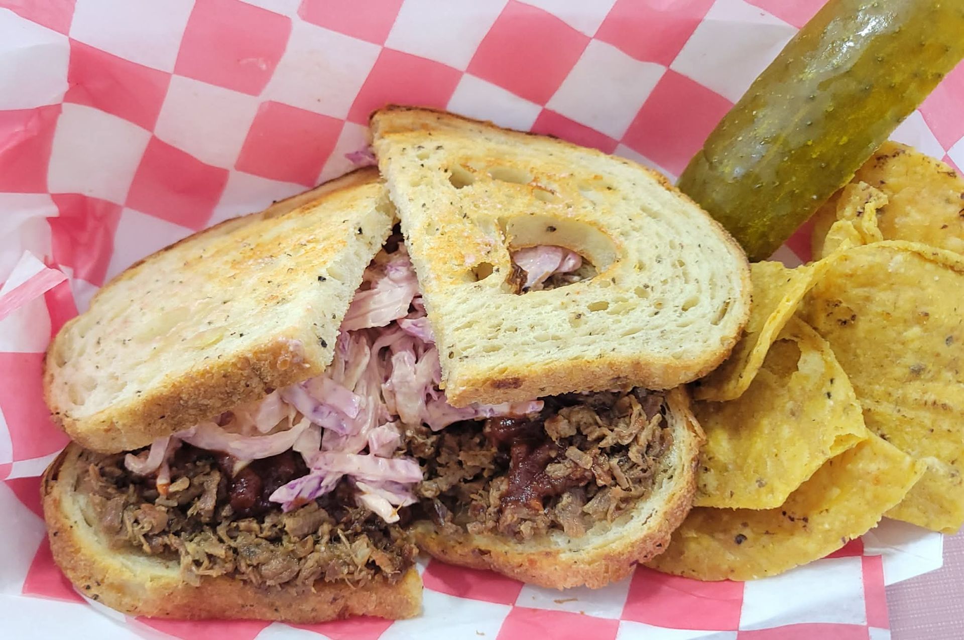 Half-cut sandwich with shredded meat and pink onions, served with tortilla chips and a pickle spear in a basket
