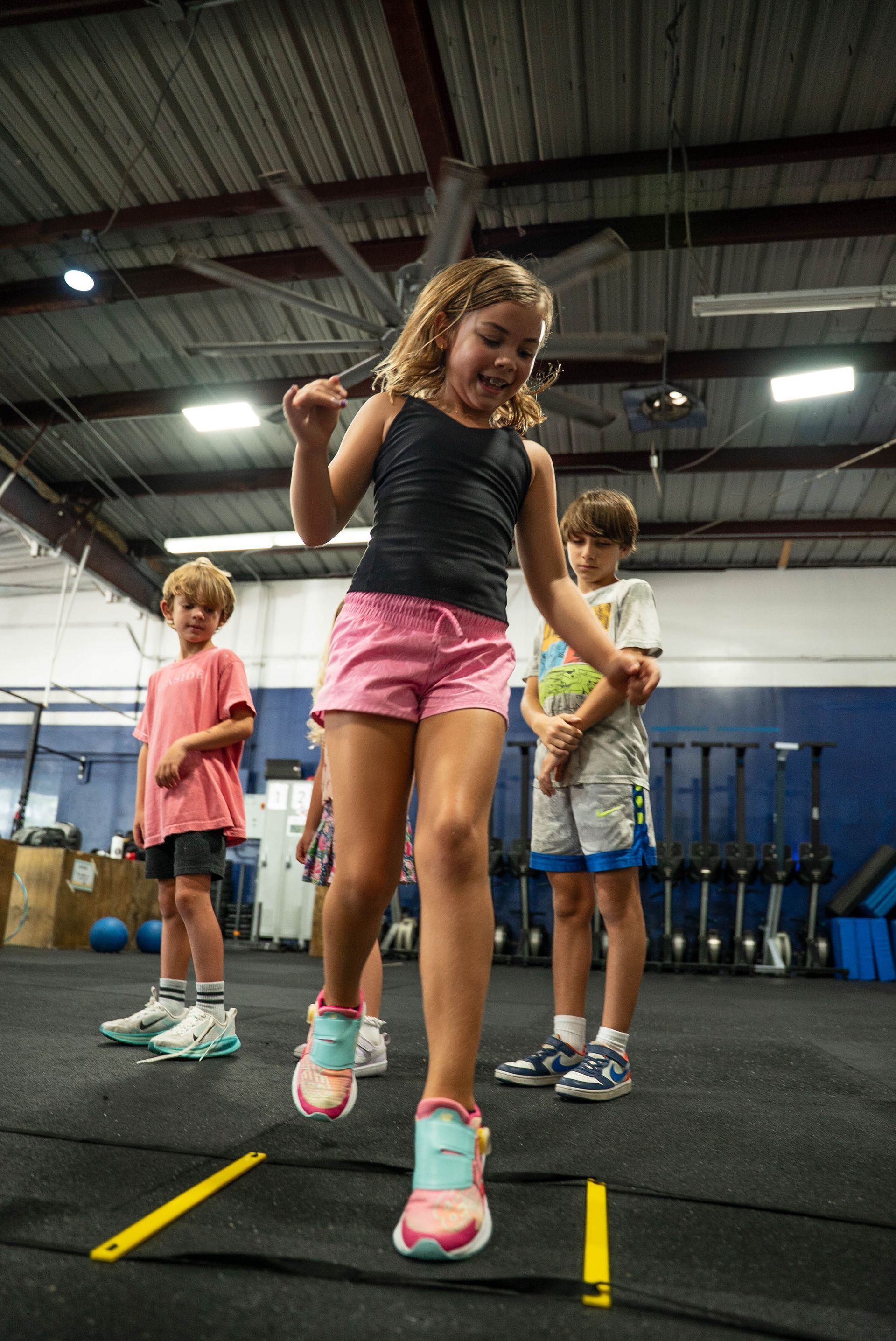 A child steps through yellow agility ladder markers on a gym floor, with two other children standing in the background.