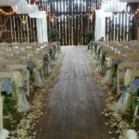 Decorated wedding aisle with white chairs, floral arrangements, and string lights in a rustic venue