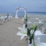 Beach wedding setup with white chairs, floral arch, and white lilies by the sea