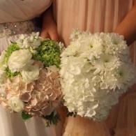 Two people holding bouquets of white and pale pink flowers with green accents, wearing peach dresses