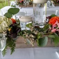 Floral table centerpiece with white candles and greenery on a wooden tray at a decorated event table