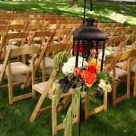 Rows of wooden chairs outdoors with a black lantern and orange-white floral arrangement