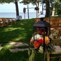 Outdoor wedding setup on a grassy lawn with lanterns, flowers, chairs, and a view of the water