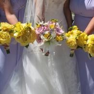 Bridesmaids holding yellow and pastel flower bouquets beside a white wedding dress
