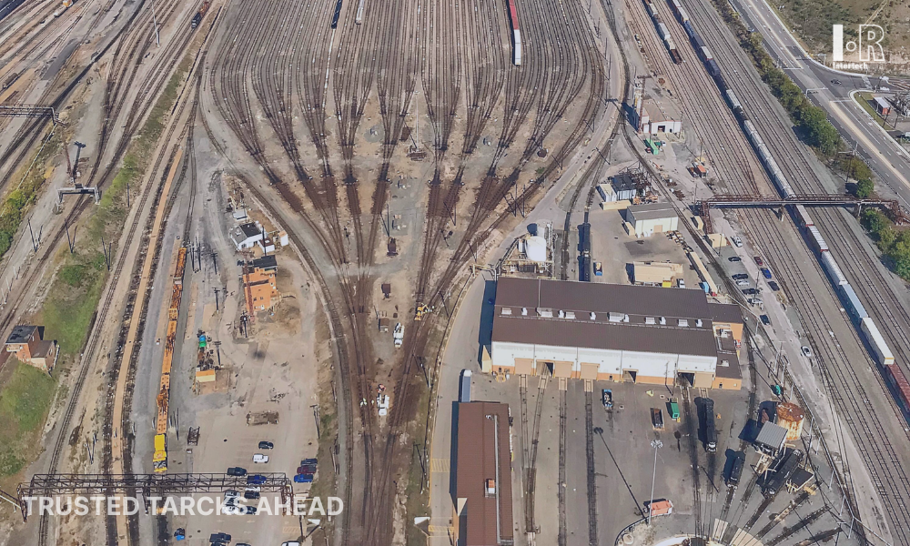 Aerial view of a rail yard with many tracks, trains, and a large warehouse beside a road.
