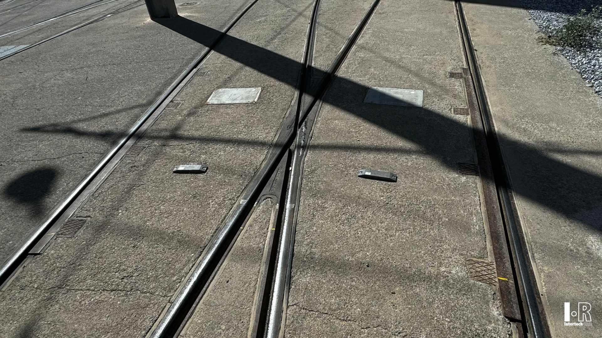 Tram tracks crossing a sunlit concrete roadway with dark shadows and lane markings. Balise System