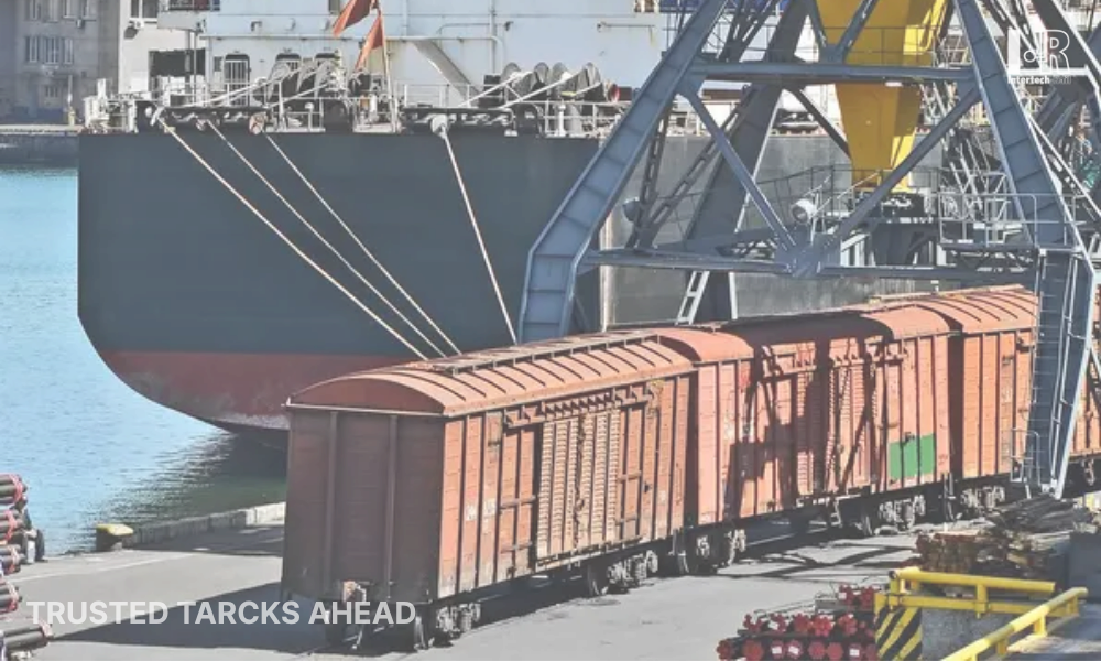 Cargo train cars on tracks beside a large ship at a port, with cranes in the background.