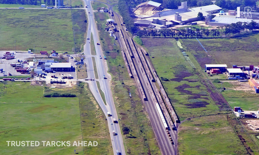 Aerial view of a road and railway tracks through green fields, with buildings nearby.