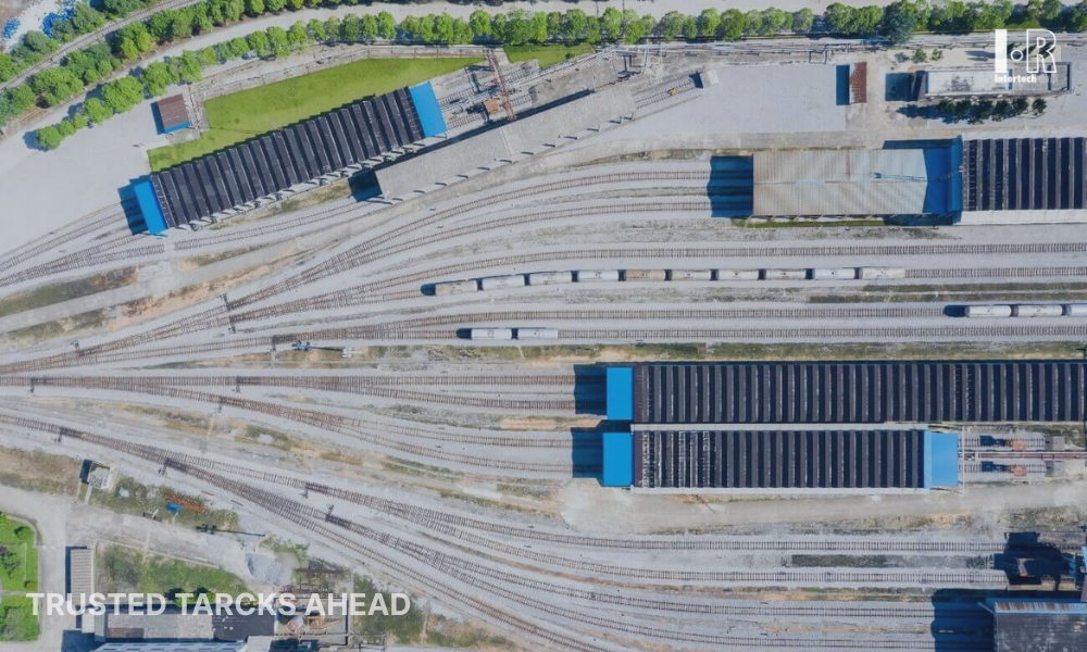 Aerial view of a rail yard with multiple tracks, freight cars, and blue-roofed structures.