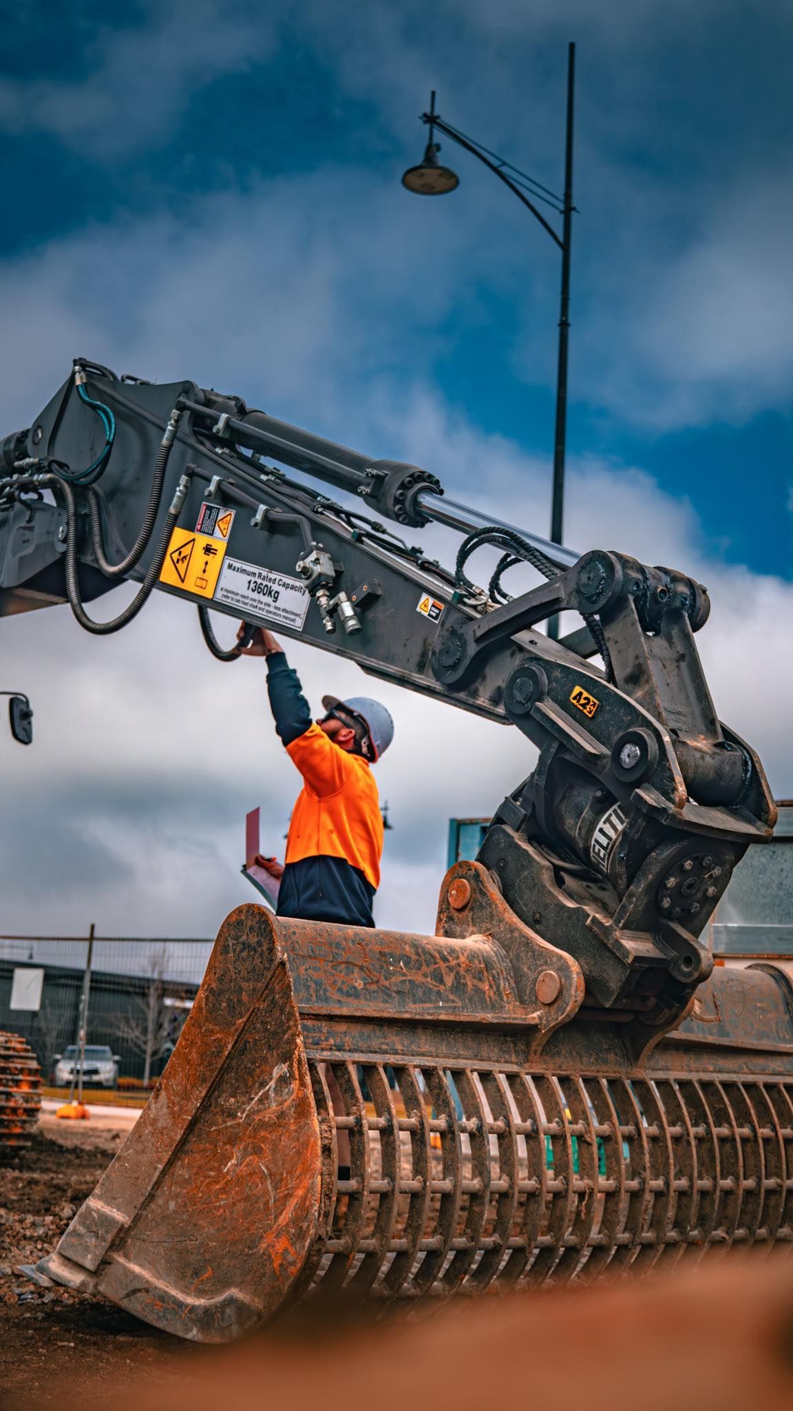 Construction worker in orange safety vest inspects excavator arm against a cloudy sky — Murphy's Salvage & Demolition in Baranduda, VIC
