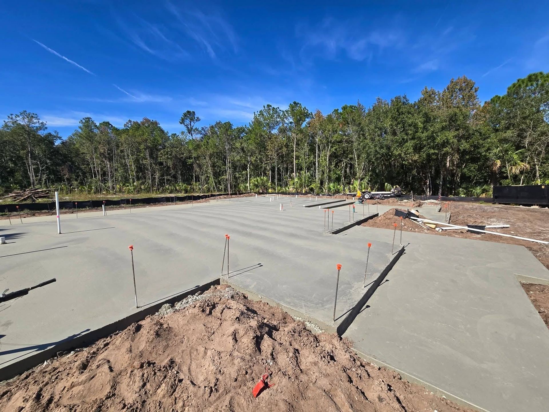 Concrete slab foundation under construction in a wooded area, with survey stakes and piles of dirt under a blue sky