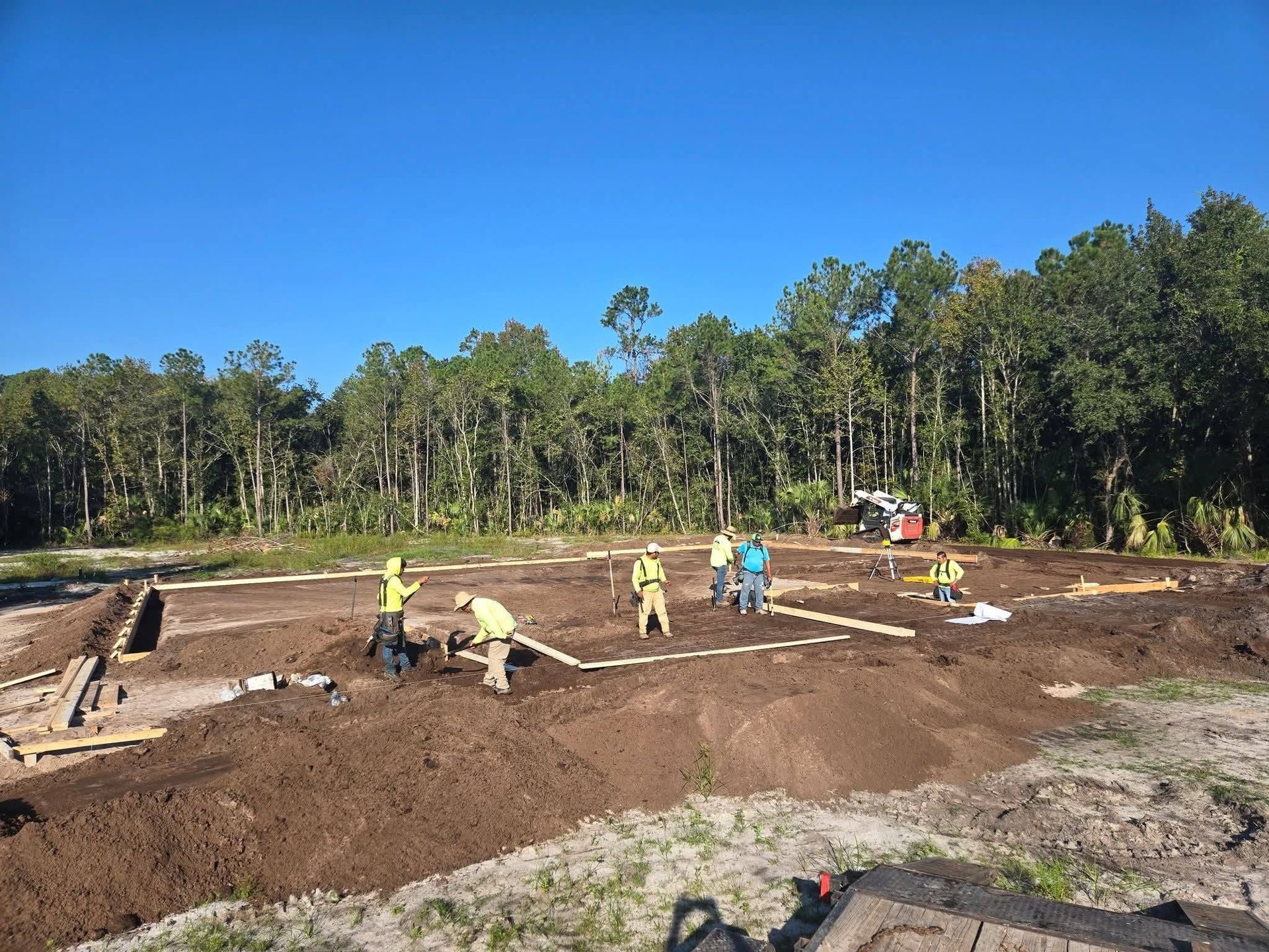 Workers in yellow shirts and hard hats spread soil at a construction site beside a forest.