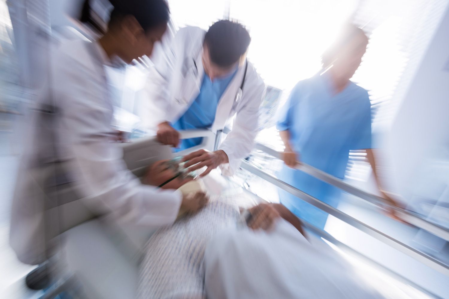 Medical staff rushing a patient on a gurney; blurred motion. Oxygen mask on patient. Hospital setting.