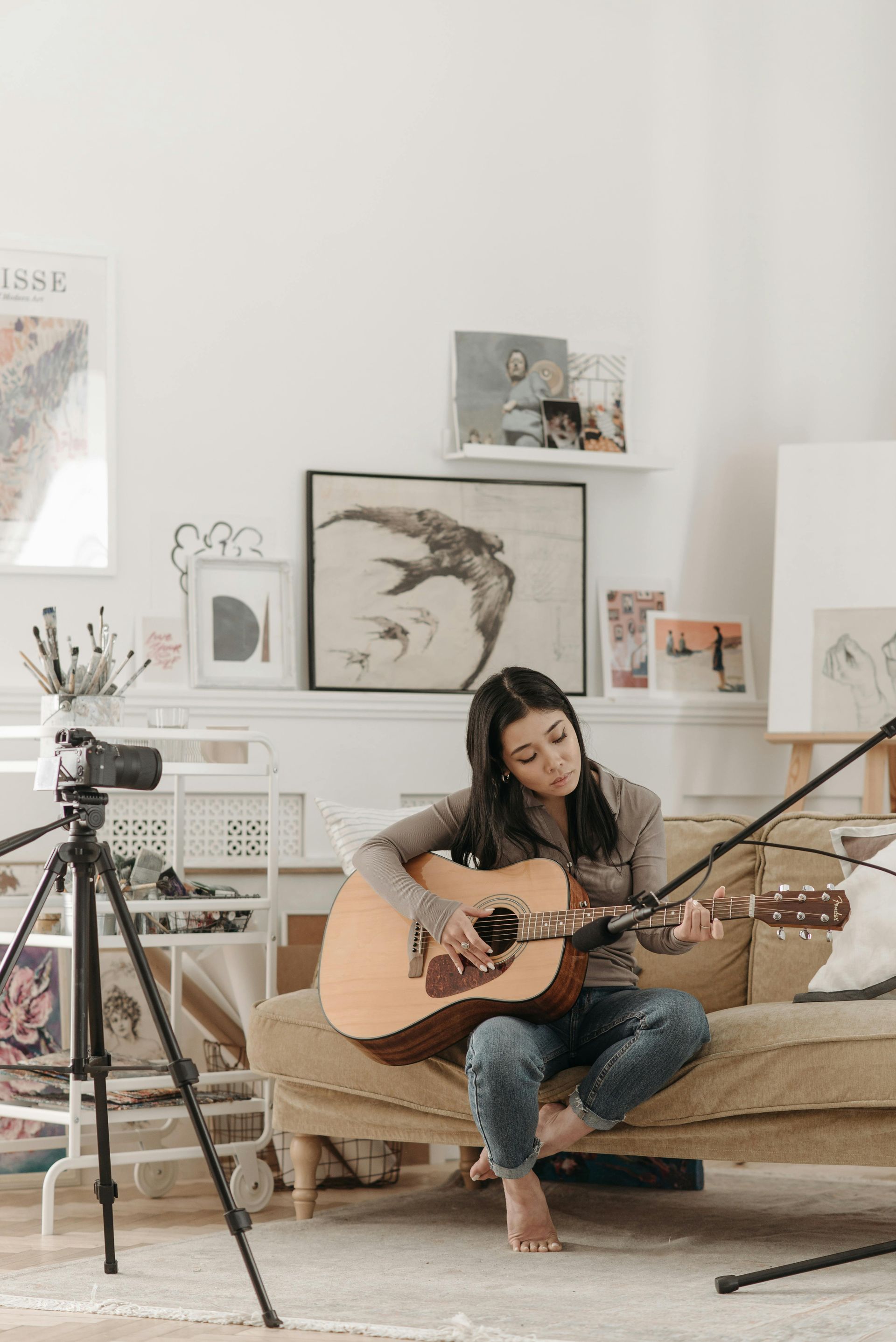 Vrouw speelt akoestische gitaar, zittend op een bank in een kunststudio, en maakt opnames met camera en microfoon.
