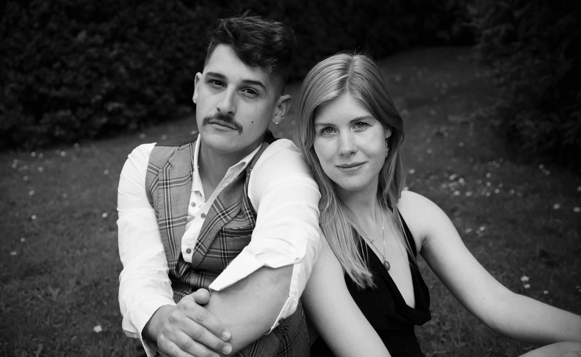 Een stel zit in het gras, de man draagt een vest met snor en de vrouw een zwarte jurk; beiden glimlachen.