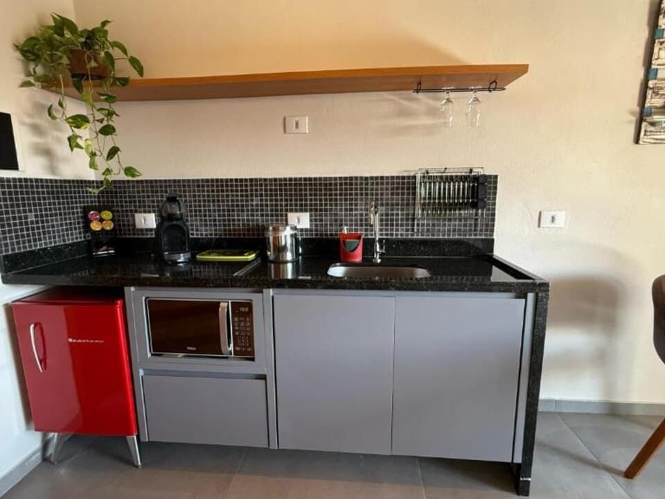 Uma pequena cozinha com um frigobar vermelho, micro-ondas embutido, armários cinza, backsplash de azulejos pretos e uma prateleira de madeira.